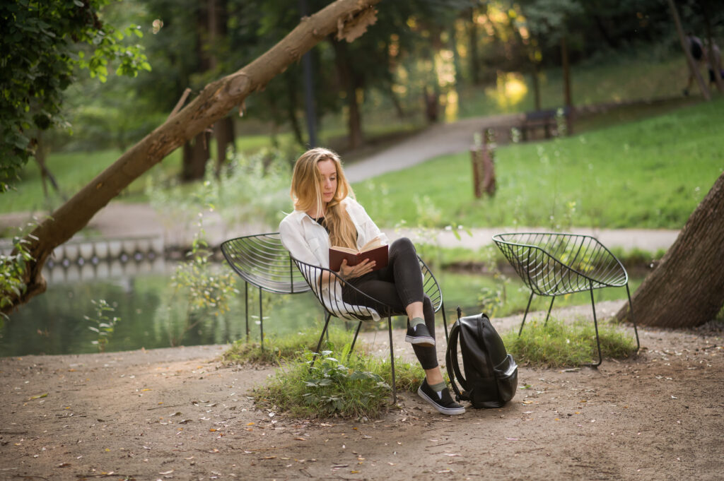Frau liest entspannt ein Buch auf einem Stuhl im Park am Wasser – Ruhe und Stressregulation bei Autoimmunerkrankungen